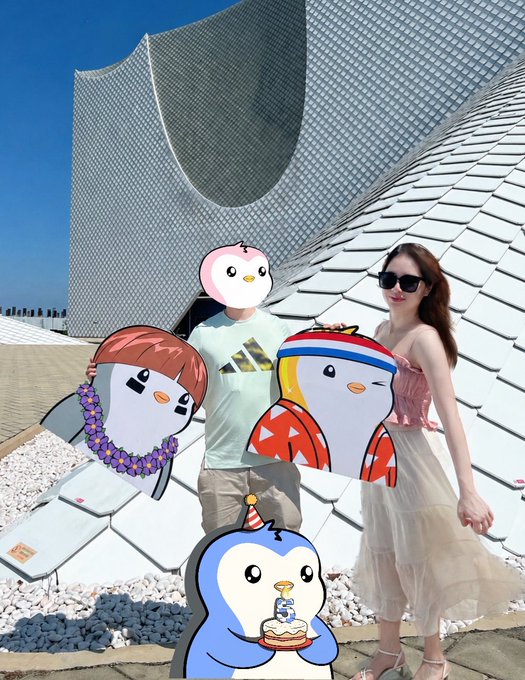 A couple poses in front of a modern architectural building, their faces covered by penguin cartoon stickers, while a small penguin sticker holding a '5' birthday cake appears in the foreground.