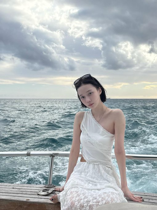 A woman in a white one-shoulder dress sits on a boat deck with the ocean and cloudy sky in the background, with sunglasses resting on her head.