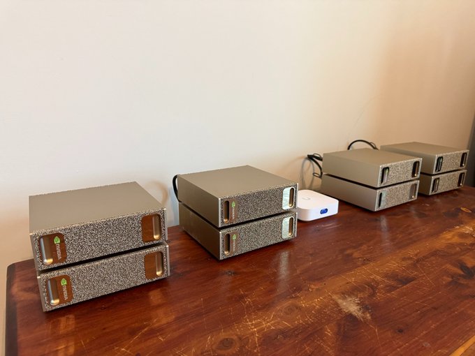A growing stack of gray AI hardware units, appearing to be NVIDIA-branded compute devices (likely Nano or Jetson-style boxes), are arranged on a wooden desk alongside a small white networking device, illustrating the expanding on-premise AI infrastructure referenced in the post.