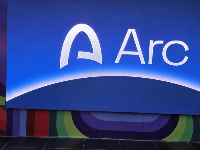 The image shows a vibrant wall display featuring the "Arc" logo—a stylized white "A" with an arching swoosh—against a bright blue background, with colorful geometric stripes in green, purple, and red along the bottom. The branding connects to the post's mention of @arc-hitects, likely referring to Arc, a tech company (Intel's Arc graphics brand) whose sleek, modern aesthetic the wall decoration represents.