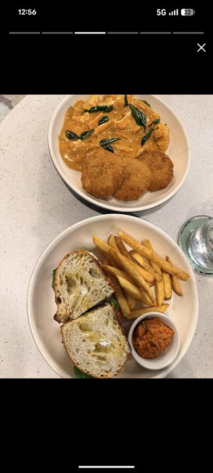 The image shows two plates of food placed side by side on a table. One plate contains fried fish with sauce, while the other has french fries accompanied by a sandwich. The photo also includes a water glass next to each dish, suggesting that they are being enjoyed as part of a meal.
