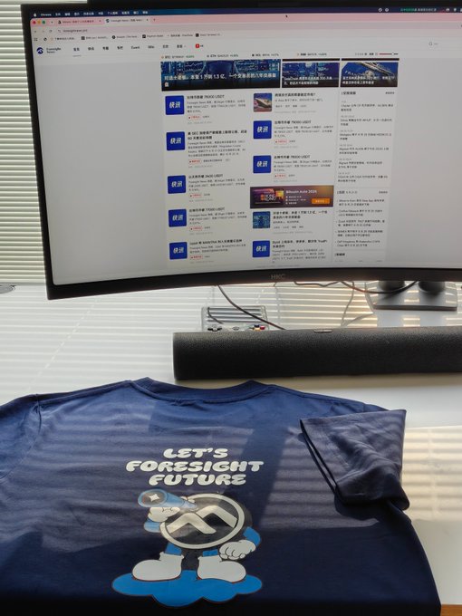 A dark blue t-shirt featuring a cartoon character and the text 'LET'S FORESIGHT FUTURE' along with a logo lies on a desk in front of a computer monitor showing a webpage with Chinese text.
