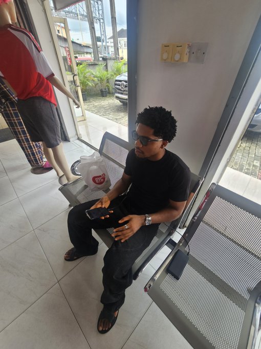 A young man wearing sunglasses and a black t-shirt sits on a metal waiting room chair, holding a smartphone, with another person's legs visible standing nearby.
