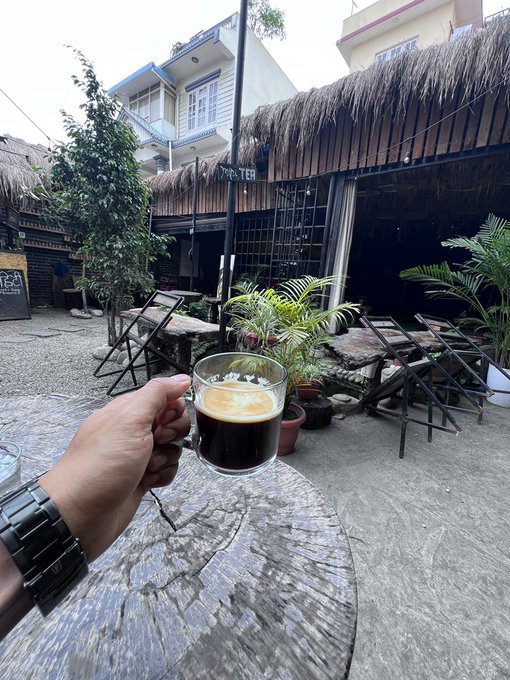 A person's hand holds a glass of espresso in the foreground, with a stone table, wooden stools, potted plants, and thatched-roof buildings visible in a courtyard setting.