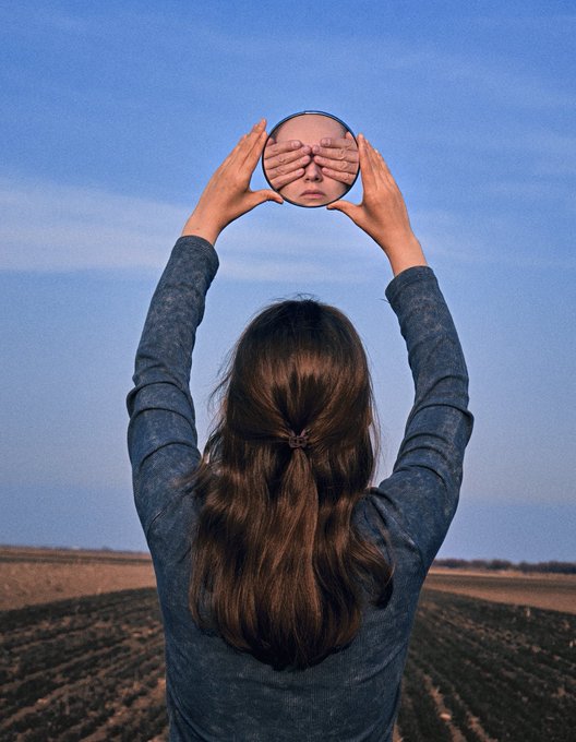 A photograph from behind of a woman with long brown hair holding a small mirror above her head, with the reflection showing hands covering a person's eyes.