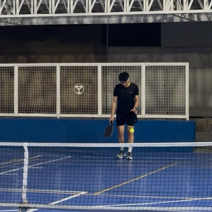 A young man in black athletic wear playing padel tennis on an indoor blue court, bouncing a yellow ball.