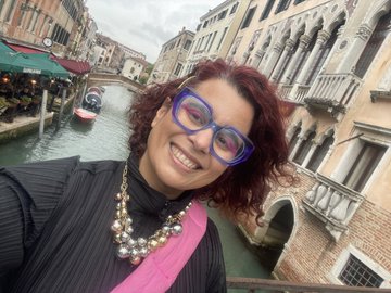 A selfie of a smiling woman with curly red hair and purple glasses, with Venetian canals and architecture visible in the background.