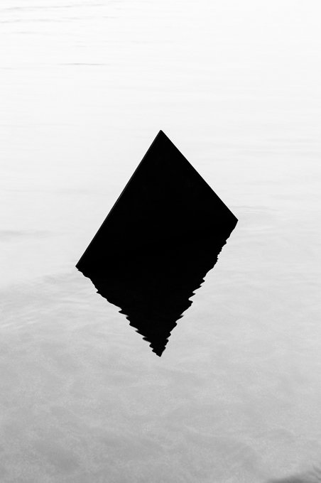 A minimalist black diamond-shaped object partially submerged in rippling gray water, with its reflection distorting downward.