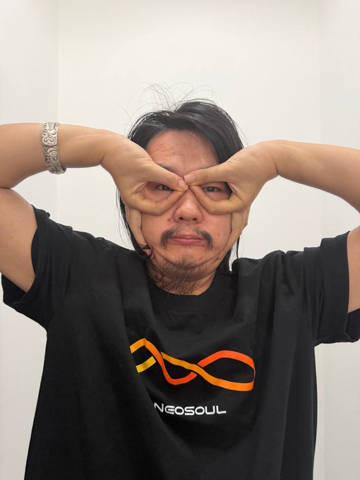 A man wearing a black 'NEOSOUL' t-shirt makes a playful gesture with his fingers, forming circles around his eyes like glasses.