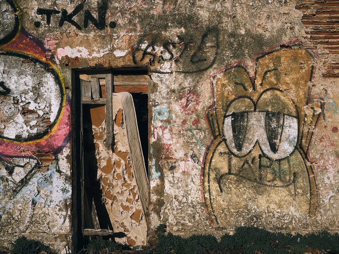 A weathered, decaying brick and plaster wall covered in graffiti, including a prominent cartoon face with large eyes and a frowning mouth, and the text 'T.K.N.' scrawled above a boarded-up doorway.