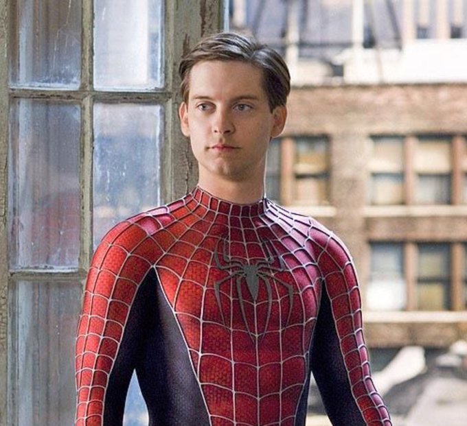 A young man (Tobey Maguire) wearing the red and blue Spider-Man suit standing in front of a window with city buildings visible in the background.
