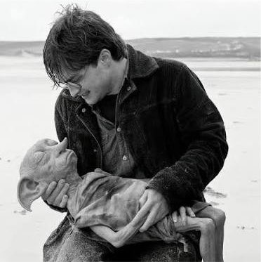 A black and white photograph shows a young boy on a sandy beach tenderly holding a strange, gaunt creature.