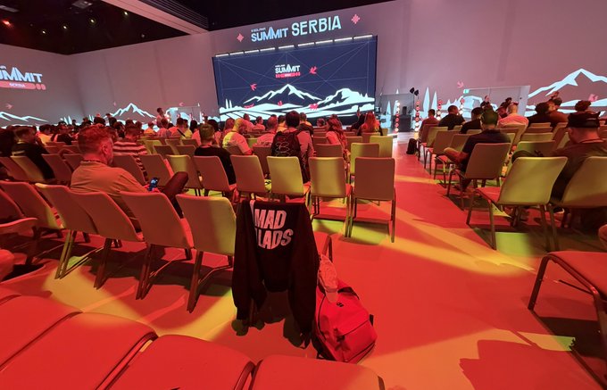 An indoor conference hall with red lighting and a stage screen displaying 'SUMMIT SERBIA', where attendees sit in rows of chairs facing the presentation.