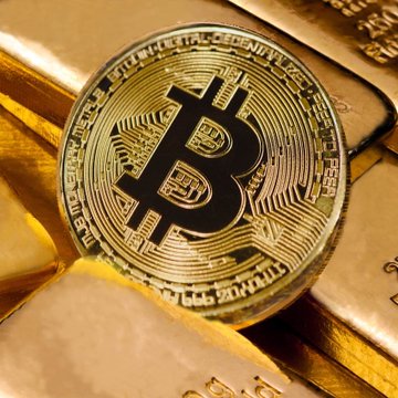 The image features a large, round, black Bitcoin coin laying next to an interesting collection of golden bars or coins with various markings. The Bitcoin coin seems to be placed near another set of gold bars stacked on top of each other. On the right side of the image, there's text that talks about how Bitcoin outperformed gold this year and what it is worth. Focus on these pieces of information in relation to the Bitcoins and gold bars in the photo.