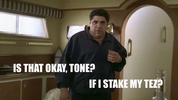 A man in a dark blue sweater stands inside an RV, with overlaid text reading "IS THAT OKAY, TONE?" and "IF I STAKE MY TEZ?" The image references Vincent Pastore's "Big P*ssy" character from The Sopranos, humorously addressing a Tezos community member nicknamed "Tone" about staking TEZ, paying tribute to the late actor.