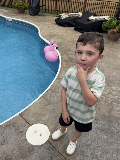 A young boy in a striped shirt stands by a swimming pool, with a pink flamingo float and a white frisbee nearby.