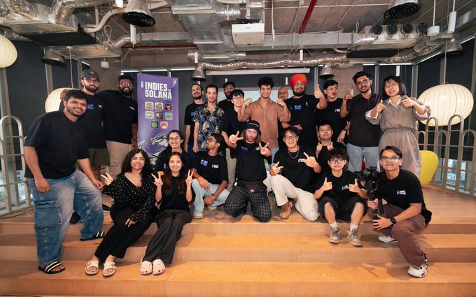 A group of people poses for a photo on wooden steps in an industrial-style space, with a banner reading 'INDIES SOLANA' visible behind them.