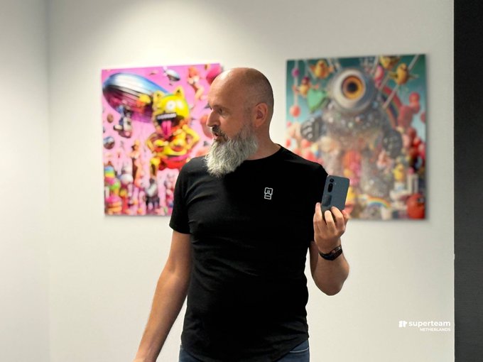 A bald man with a long white beard wearing a black t-shirt holds up a blue smartphone in front of colorful NFT artwork in what appears to be a gallery setting.