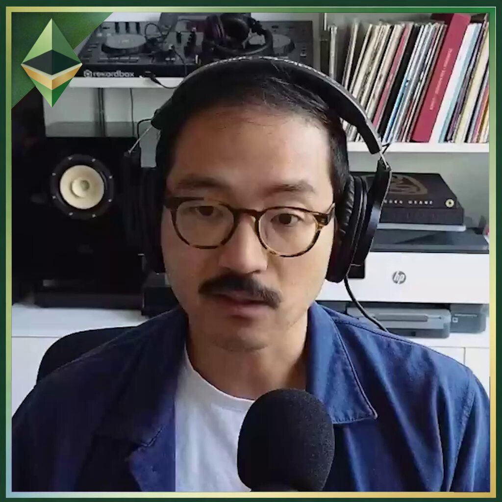 The image features a man wearing headphones and glasses sitting at a desk with a microphone in front of him. He appears to be engaged in a conversation or interview, as indicated by his facial expression. The background includes several bookshelves filled with various books, suggesting that he might be discussing the world of cryptocurrencies or related topics.