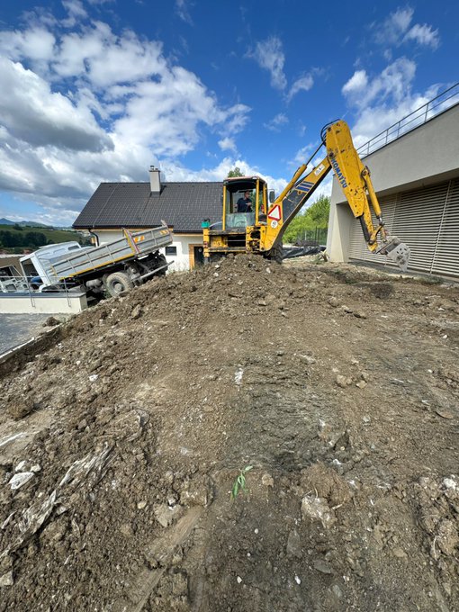 A yellow excavator and a dump truck are working on a dirt construction site next to a house and a building with a roll-up door under a partly cloudy sky.