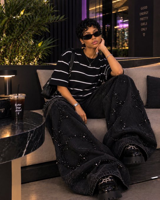 A woman with short curly hair and dark sunglasses lounges on a gray couch wearing a black and white striped top and embellished wide-leg black jeans with chunky black shoes, with "GOOD ENERGY PRETTY GIRLS ONLY ♡" visible on a sign in the background.