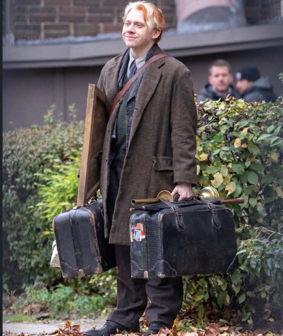 A young man with reddish-blonde hair wearing a long brown coat, holding vintage suitcases, a framed picture, and brass horns, standing outside near a hedge.
