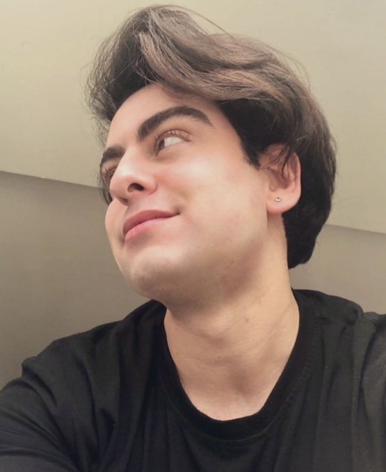 A close-up selfie of a young man with dark wavy hair, wearing a black t-shirt and an earring, looking upward with a slight smile.