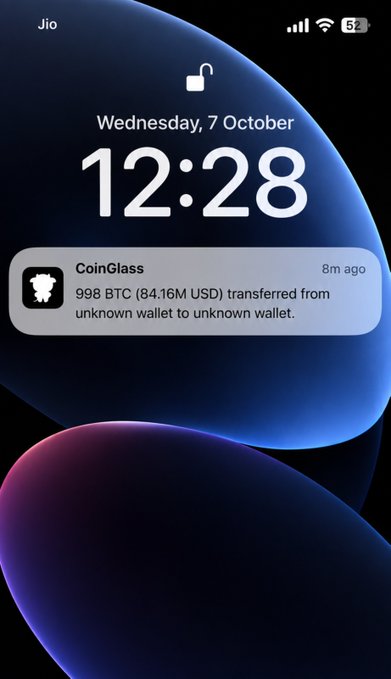 iPhone lock screen on Wednesday, 7 October at 12:28 showing a CoinGlass alert: 998 BTC (84.16M USD) transferred from unknown wallet to unknown wallet.