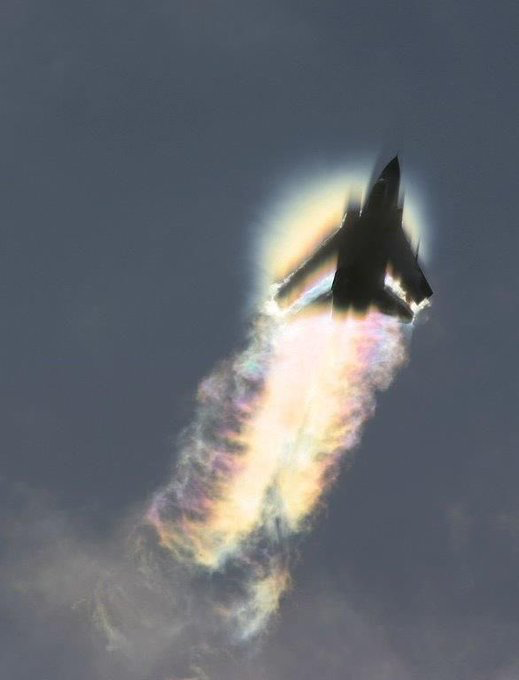A fighter jet is captured mid-flight from below, surrounded by a luminous vapor cone with rainbow colors forming in its exhaust trail against a gray sky.