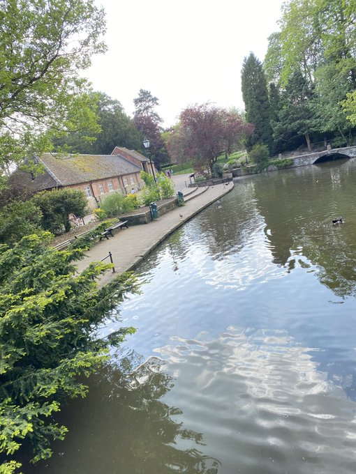 A serene view of a canal or river lined with lush green trees and a stone walkway, with a brick cottage and a stone bridge visible in the background.