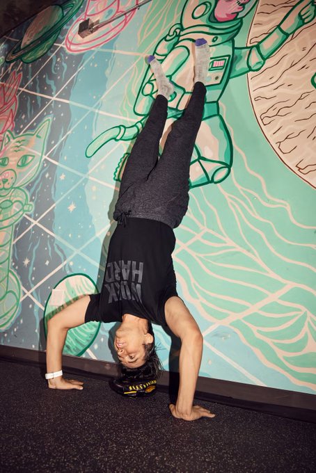 A person performing a headstand against a colorful space-themed mural, wearing a black tank top that reads 'MONK MODE' (inverted).