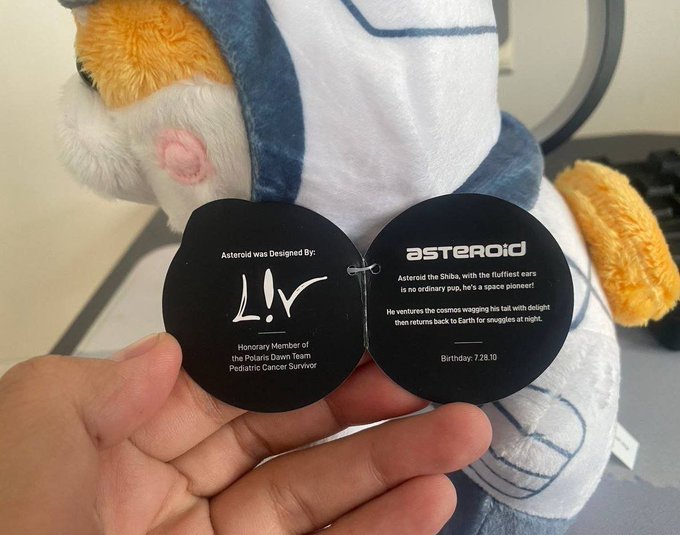 A hand holds open a black heart-shaped tag attached to the "Asteroid" plush toy, designed by "Liv," an Honorary Member of the Polaris Dawn Team and Pediatric Cancer Survivor, with a birthday of 7.28.10, tying into the post's mention of Asteroid's anniversary and Liv's birthday.