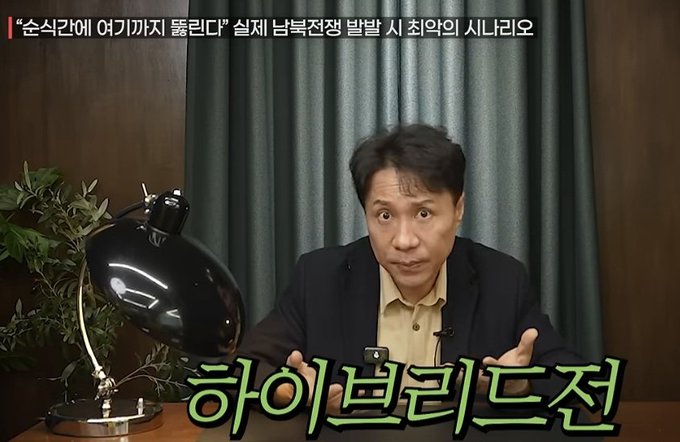 The image shows a man in a dark suit speaking directly to the camera, seated at a desk with a black lamp and a smartphone, with bold green Korean text overlay reading "하이브리드전" (Hybrid Warfare) and a top banner stating "순식간에 여기까지 뚫린다면 실제 남북전쟁 발발 시 최악의 시나리오" (Worst case scenario if the South is breached this quickly during actual Korean War outbreak). The content aligns with the post's discussion of North Korea's hybrid warfare strategy, combining cyber attacks like communications and GPS disruption with physical military strikes.
