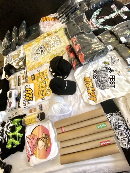 A table covered with merchandise including t-shirts, hats, patches, and TubeRex rolling paper tubes, featuring designs related to "Vape Fest" and "OPER" brands.