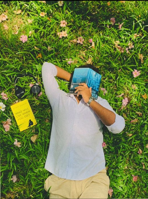 A person lies on grass scattered with pink flowers, reading a blue book over their face, with sunglasses and a yellow book titled 'Who Will Cry When You Die?' resting beside them.