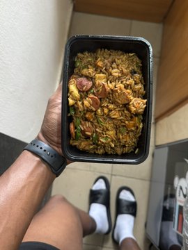 A person wearing a black smartwatch and white socks holds up a black takeout container filled with a savory rice dish featuring sausage, vegetables, and herbs, illustrating the post's sentiment that "good meals saves lives." The overhead shot captures the hearty home-cooked or restaurant-style meal from a casual, personal perspective.