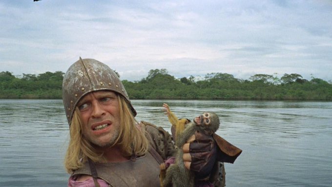 A scene from a film showing a man with long blonde hair wearing a rusty metal helmet, holding a small monkey while standing in front of a river and jungle.