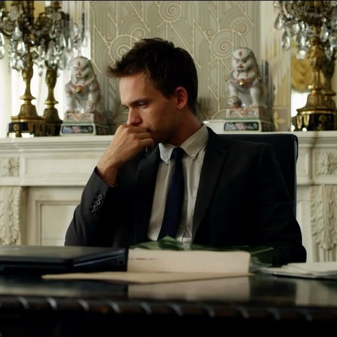 A man in a dark suit and tie sits thoughtfully at a desk in an ornately decorated room with decorative statues and chandeliers.