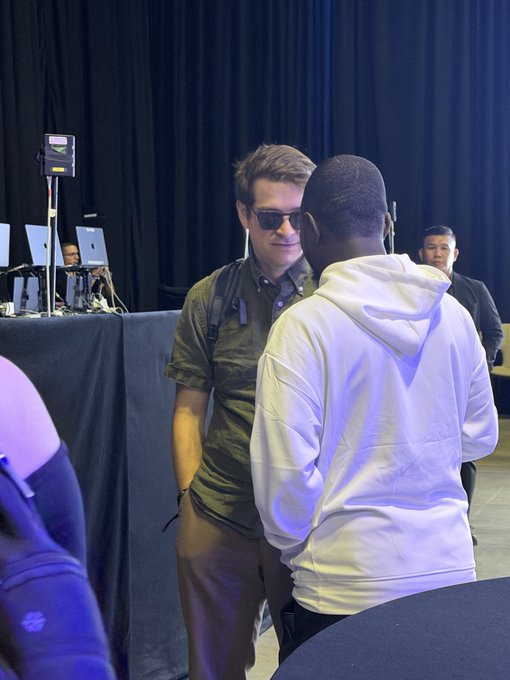 Two men converse at what appears to be a tech event, with one wearing sunglasses and a green shirt and the other in a white hoodie, while laptops and crew equipment are visible in the background.