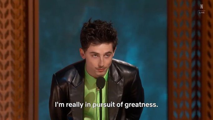 A young man in a black leather jacket and green shirt speaks at a microphone, with subtitles reading 'I'm really in pursuit of greatness.'