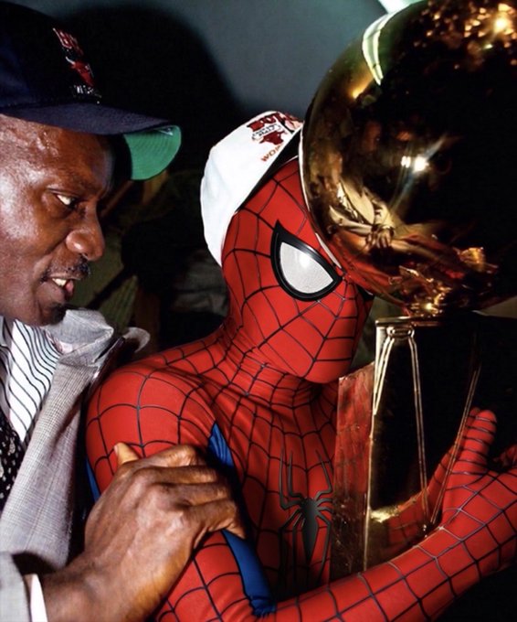 A man in a Bulls cap and gray suit embraces someone in a Spider-Man costume who is holding a large golden championship trophy.