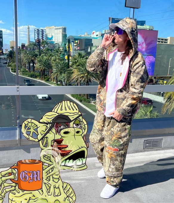 A person wearing a camouflage tracksuit and reflective sunglasses stands on a balcony overlooking the Las Vegas Strip, with an overlaid cartoon illustration of a reptilian character holding a 'GM' mug.