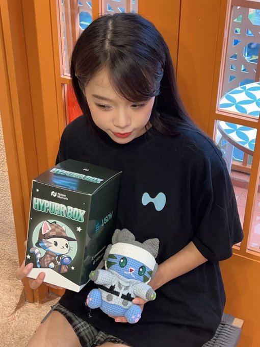 A young woman wearing a black t-shirt holding a 'HYPURR BOX' NFT collectible box alongside a crocheted cat plushie doll.
