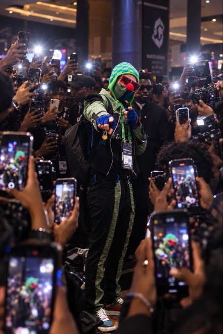 A person in a colorful costume with a green hood, sunglasses, and a red clown nose, surrounded by a crowd of fans taking photos with their smartphones.