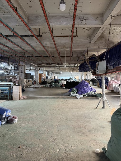 An empty, abandoned industrial warehouse or factory interior with exposed ceiling beams, scattered debris, tarps, and piles of materials on a concrete floor.