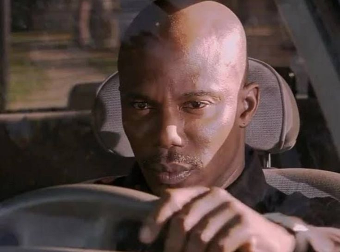 Close-up shot of a bald man driving a car, looking intently forward with his hand on the steering wheel.
