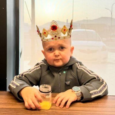A baby wearing a large jeweled crown, a gray track jacket, and a wristwatch, sitting at a wooden table with a small jar of orange juice.