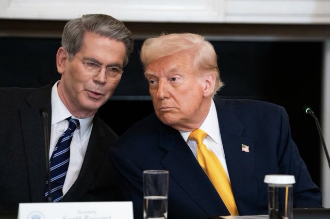 The image shows two men sitting at a table, one wearing a suit and tie with glasses while the other has a yellow tie. They are both looking at something off-camera, possibly engaged in a conversation or observing an event. The caption "Is Trump good or bad for $XRP" suggests that there is some discussion about Trump's impact on cryptocurrency markets, specifically Ripple, which could be related to their current situation.