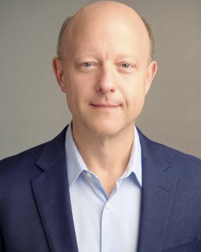 A headshot of a bald man in a blue blazer and light blue shirt against a gray background.