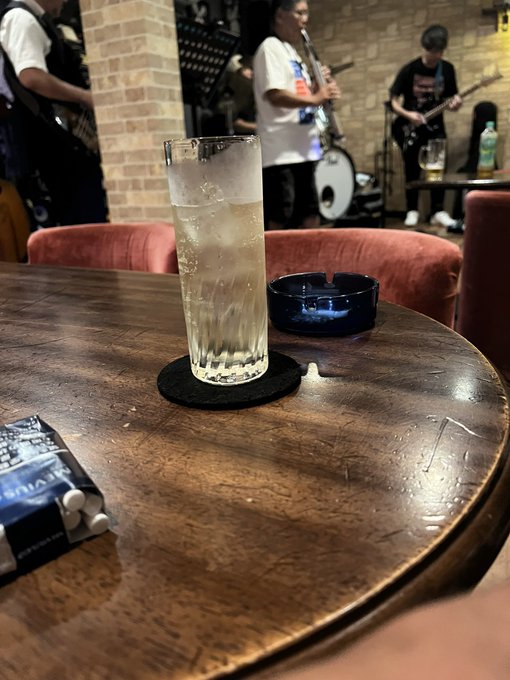 Photo of a tall glass of water with ice and a blue glass ashtray on a wooden table, with a band playing guitars visible in the background at what appears to be a live music venue.